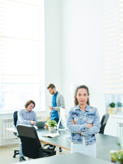 Serious successful female startup leader with crossed arms looking at camera and standing by table in meeting room